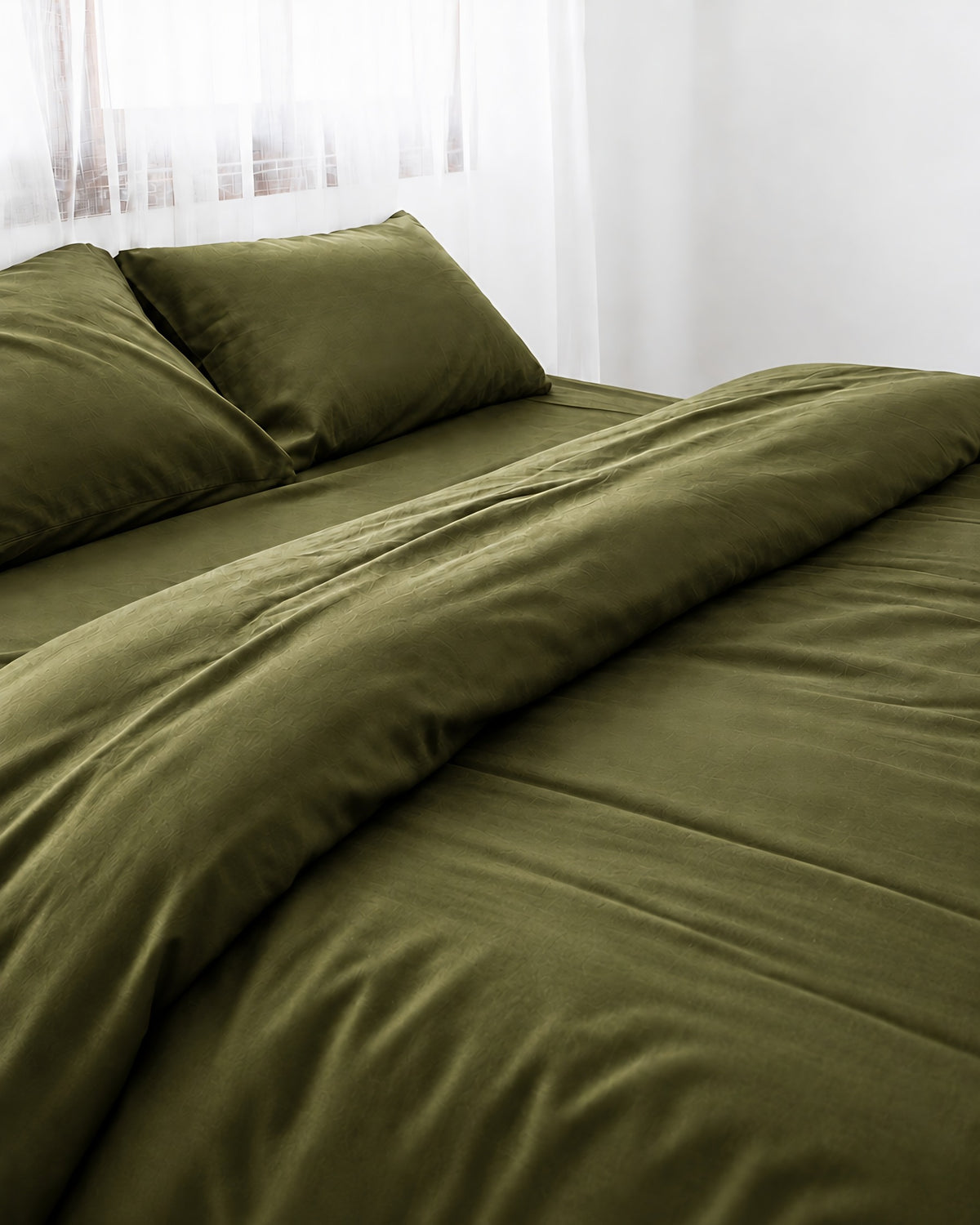 Juniper Green silky cotton duvet cover with one corner turned back on a bed by a white sheer curtain