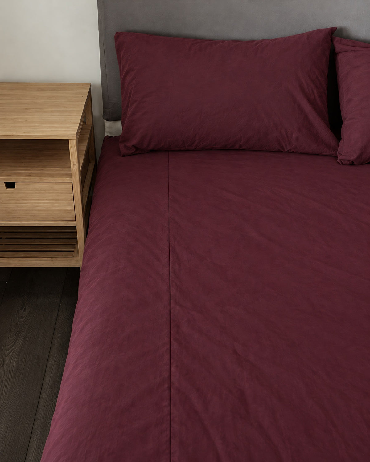 Burgundy Red plain cotton fitted sheet and pillow on a bed beside an oak bedside table
