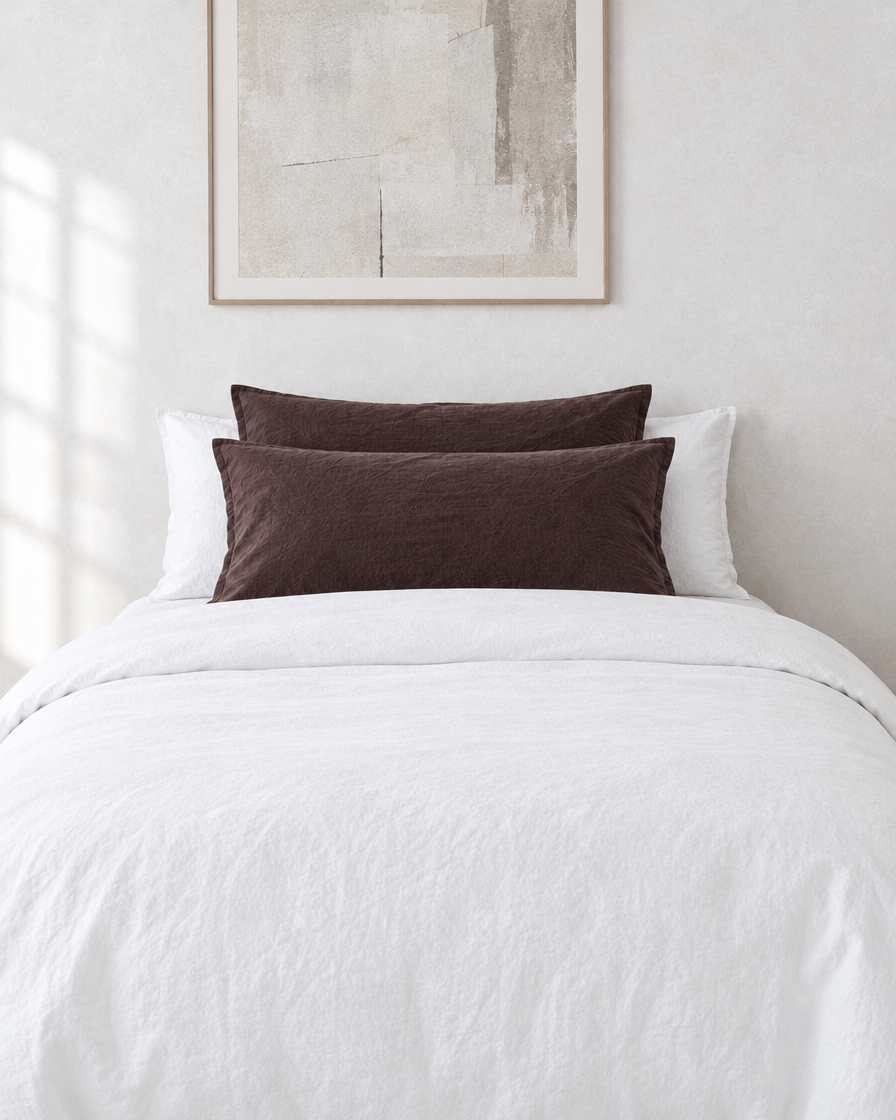 Chocolate plain cotton pillowcases on pillows against a white bed between bedside lamps.