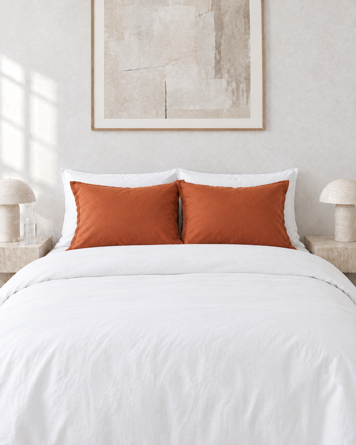 A pair of standard Terracotta plain cotton pillowcases in front of white pillows, between travertine bedside lamps.