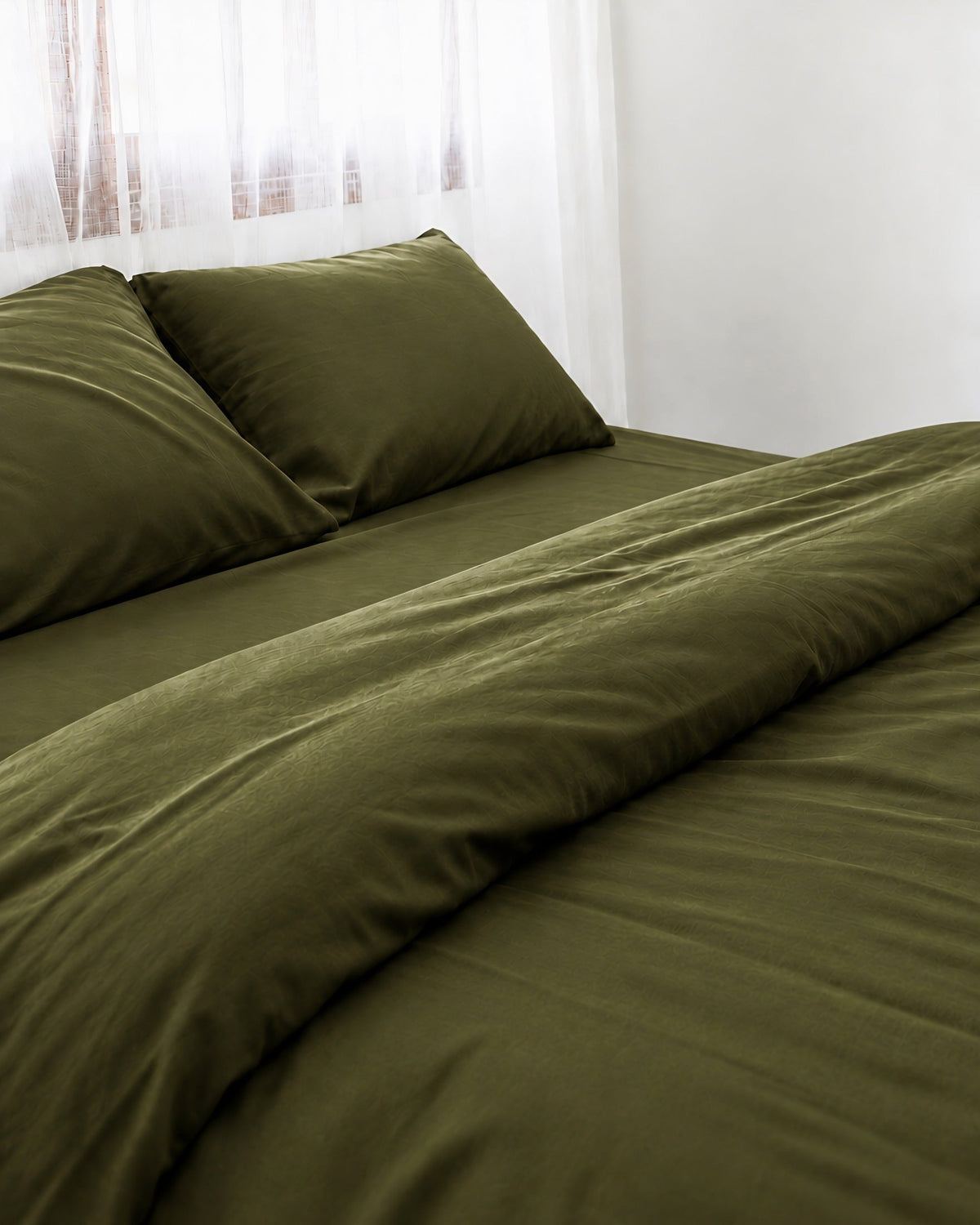 Two pillows in Juniper Green silky cotton pillowcases at the head of a made bed by a sheer curtain
