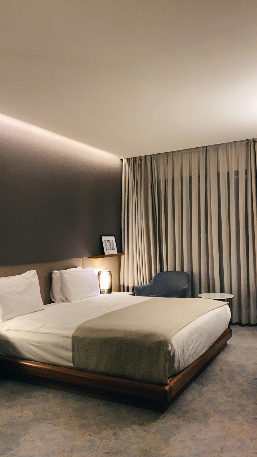 Hotel bedroom with a made bed in white bedding, a runner folded across the foot and full-height curtains behind.