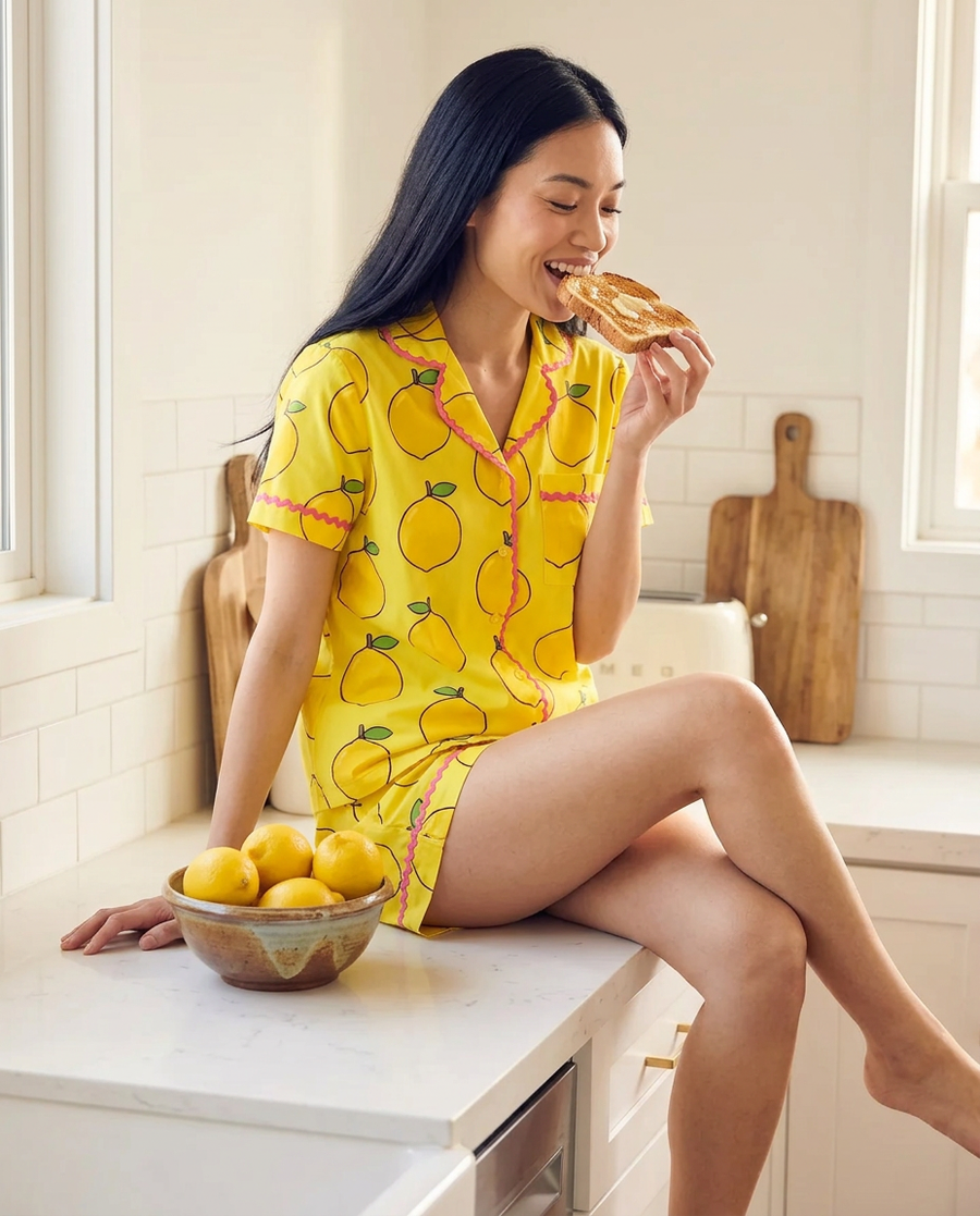 A woman in the Ripe Lemon short pyjama set sitting on a kitchen counter beside a bowl of lemons.