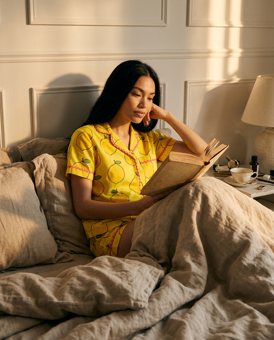 A woman in the Ripe Lemon short pyjama set reading in a bed of rumpled natural bedding.