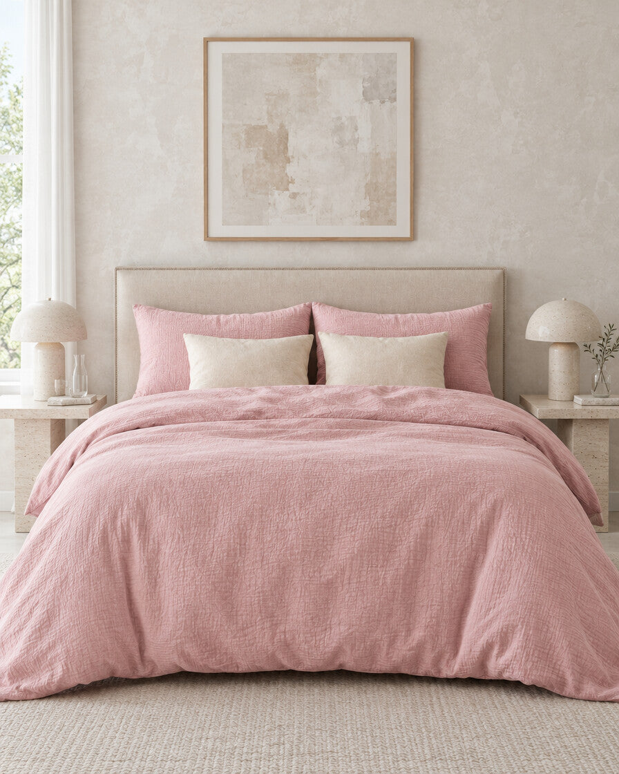 Dusty pink cloud cotton bedding set against a limewashed plaster wall between travertine bedside tables