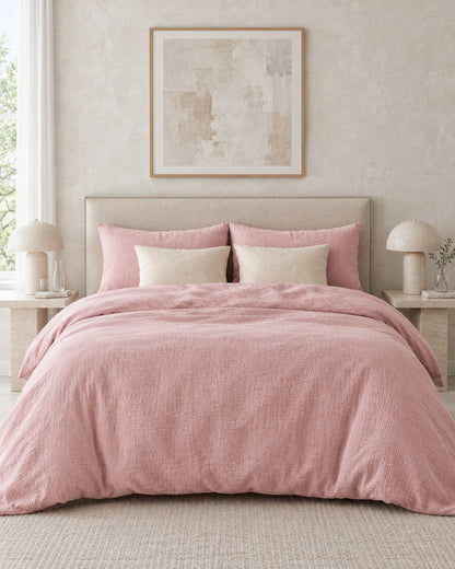 Dusty pink cloud cotton bedding set against a limewashed plaster wall between travertine bedside tables