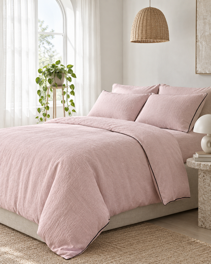 Pale Pink piped cloud cotton bedding with fine dark piping beside an arched window, a trailing pothos and rattan pendant.