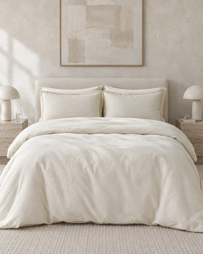 Cream piped cloud cotton bedding set against a limewashed plaster wall between travertine bedside tables