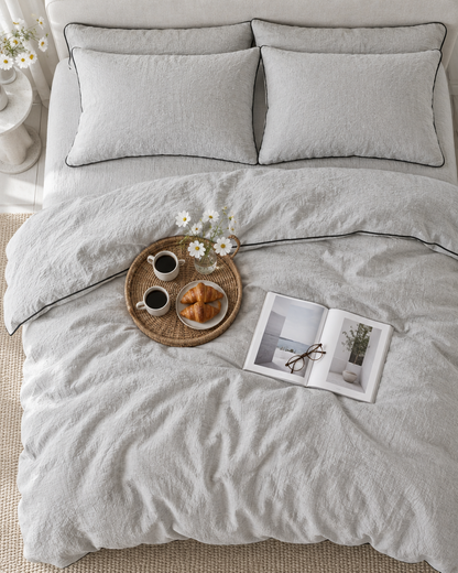 Warm Grey piped cloud cotton bedding from overhead, black piping on pillows, a rattan breakfast tray and reading glasses.