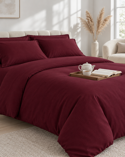 Burgundy Red plain cotton bedding with a tea tray and open book at the foot, a boucle armchair and dried pampas behind.