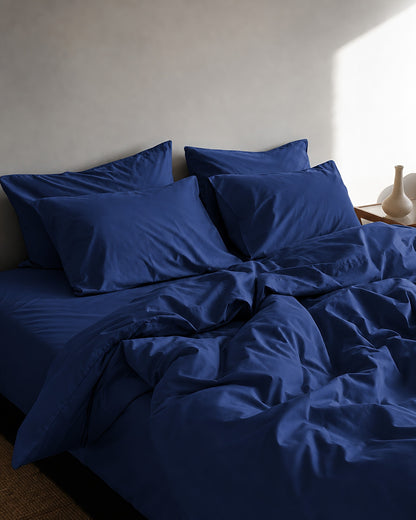 Marine Blue silky cotton bedding with a rumpled duvet and stacked pillows beside a ceramic bud vase