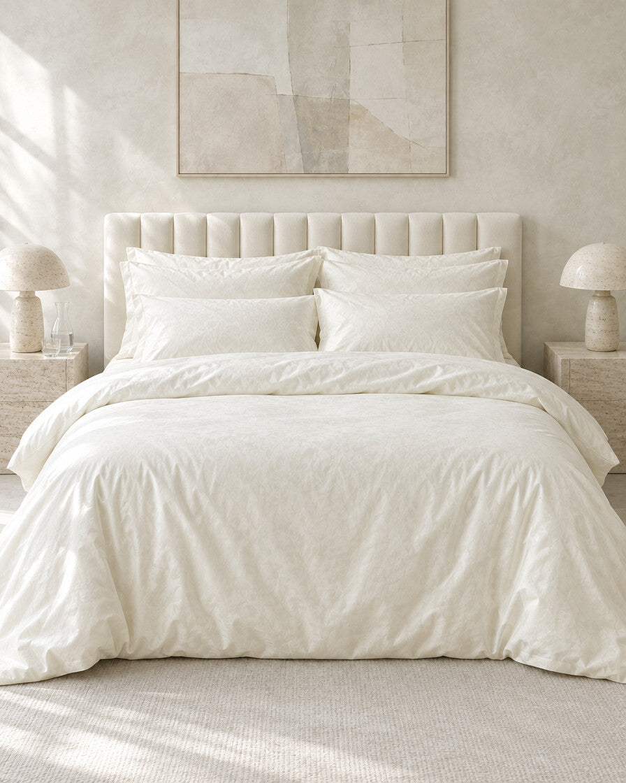 Ivory silky cotton bedding set against a limewashed plaster wall between travertine bedside tables