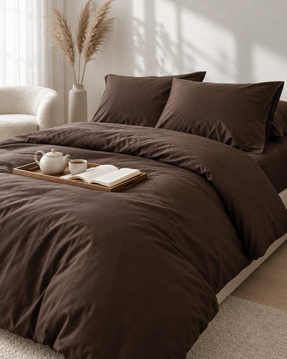 Chocolate brown silky cotton bedding set with a tea tray and open book, a boucle armchair in the corner