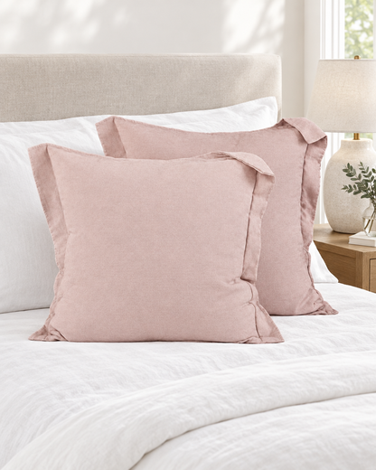 Two solid blush lyocell-linen flanged cushion covers on a made bed with white linen and a ceramic lamp