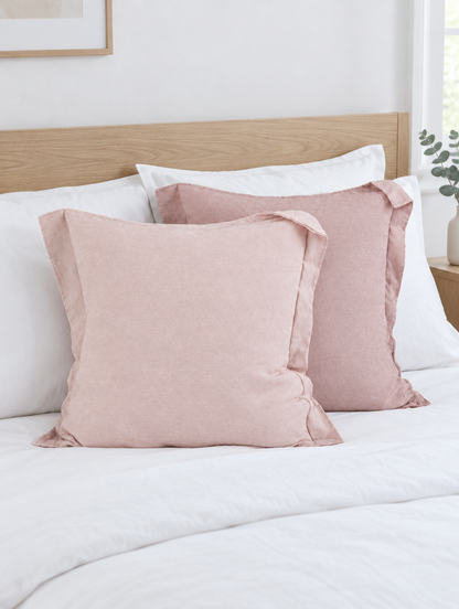 Two blush pink two-tone cushions propped on white cotton bedding below an oak headboard