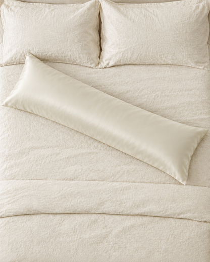 Overhead view of a Vanilla body pillow in a cream silky cotton sateen pillowcase lying across a cream bed.