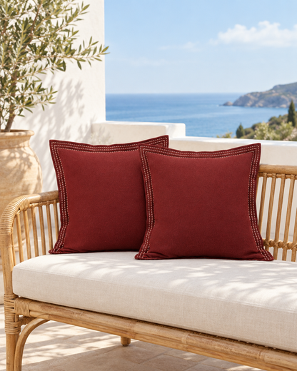 Two burgundy ramie cotton cushions with ivory stitched borders on a rattan sofa on a sea-view terrace