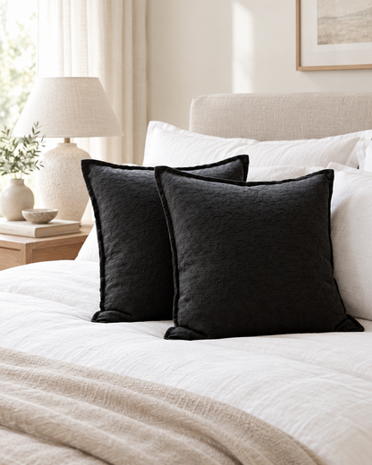 Charcoal flanged jacquard cushions on a made bed with white linen and a bedside lamp