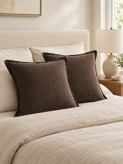 Chocolate check jacquard cushions with black whipstitch edges on a linen duvet beside a ceramic lamp