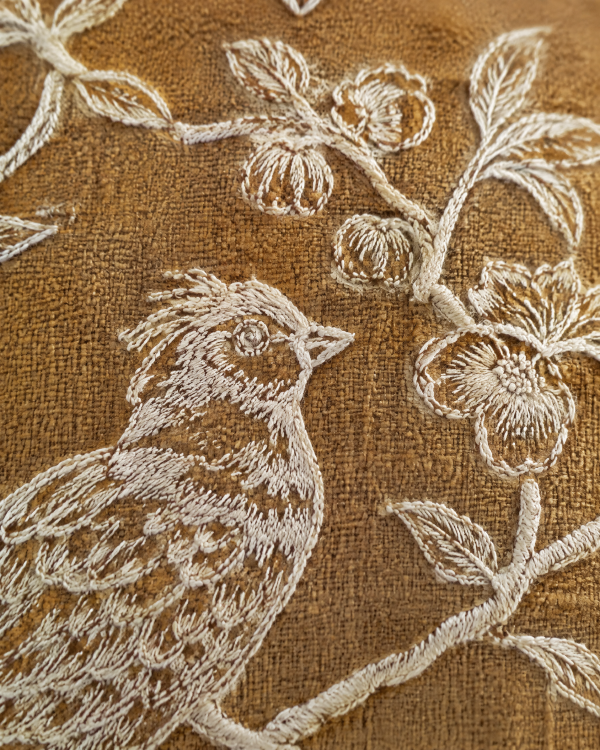 Macro close-up of an ivory embroidered bird and blossom branch on matte mustard ramie