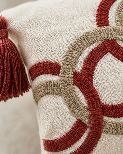Macro close-up of the ring embroidery, brick red and khaki chain-stitch rings crossing on ivory cloth, with a red corner tassel