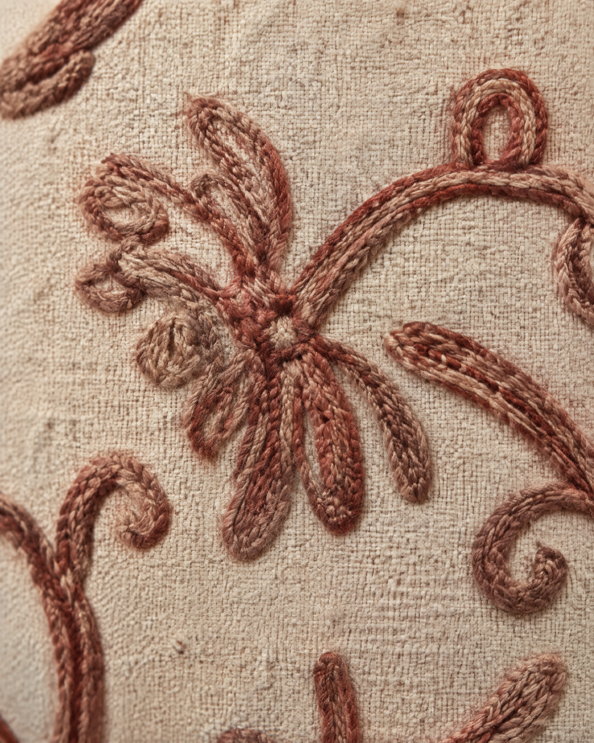 Macro close-up of the vine embroidery, thick rust-rose crewel vines and an open flower spray on slubby sand linen