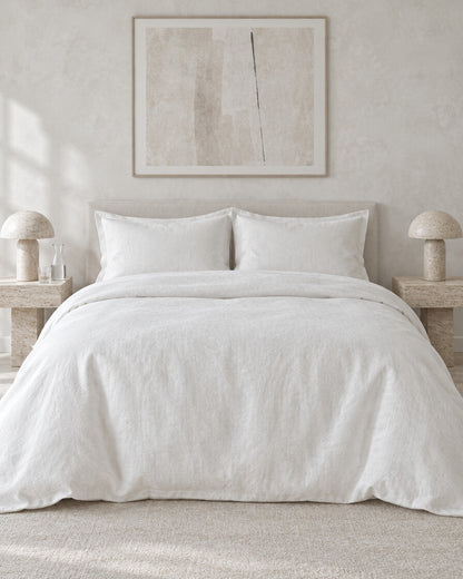 White French linen duvet cover against a limewashed plaster wall between travertine bedside tables