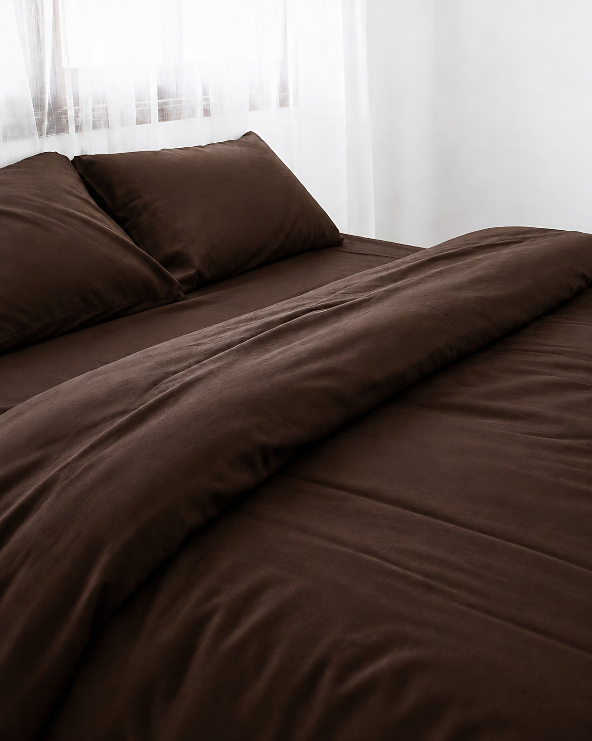 Chocolate silky cotton duvet cover with one corner turned back on a bed by a white sheer curtain
