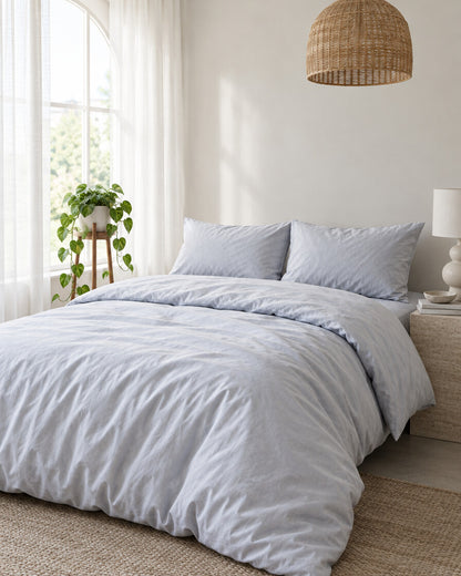 Pale blue-grey silky cotton duvet cover beneath an arched window with a trailing pothos and rattan pendant