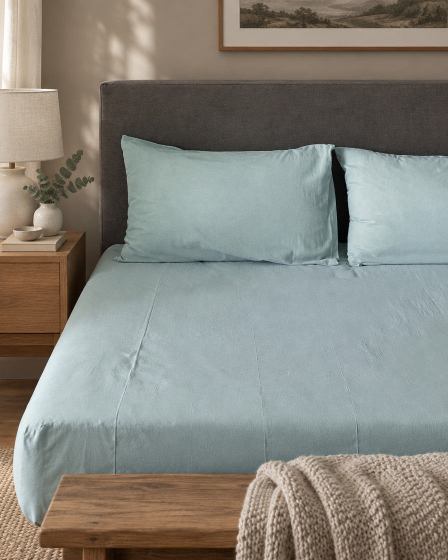 Pale aqua plain cotton fitted sheet and pillowcases against a grey headboard, with a wooden bench