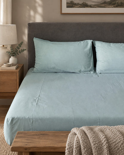 Pale aqua plain cotton fitted sheet and pillowcases against a grey headboard, with a wooden bench