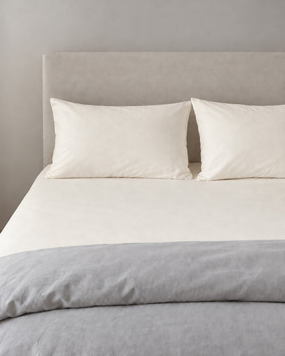 Off White plain cotton fitted sheet and pillowcases in warm ivory, grey duvet folded at the foot