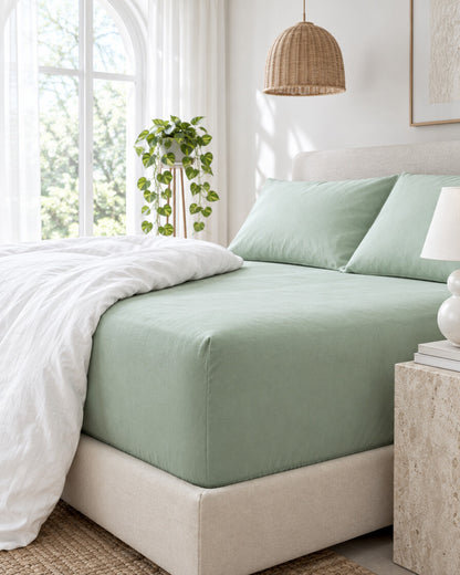Sage green plain cotton fitted sheet beneath an arched window with a trailing pothos and rattan pendant