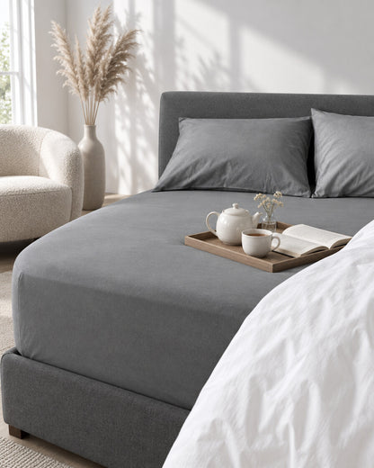 Grey plain cotton fitted sheet with a tea tray and open book, a boucle armchair in the corner
