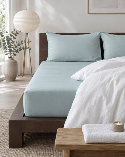 Pale blue plain cotton fitted sheet in a calm Japandi bedroom on a low dark-oak bed