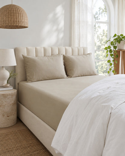Sand plain cotton fitted sheet beneath an arched window with a trailing pothos and rattan pendant