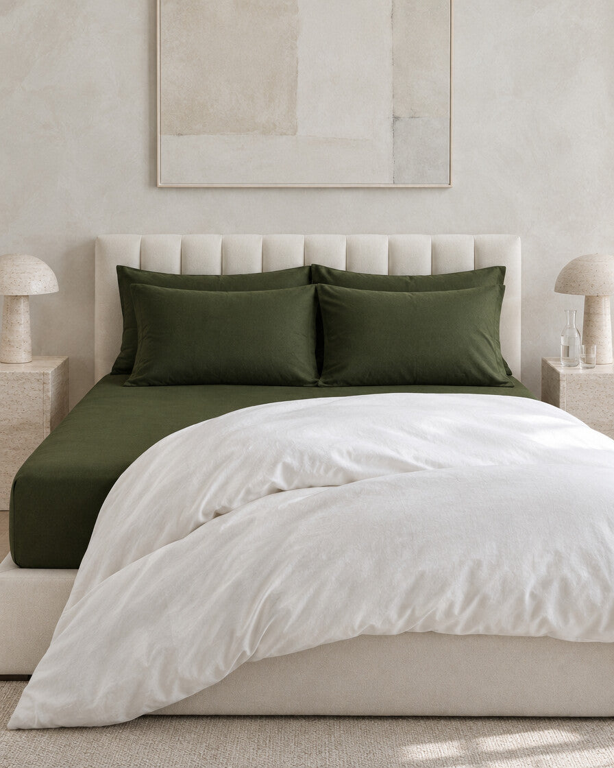 Forest green plain cotton fitted sheet against a limewashed plaster wall between travertine bedside tables