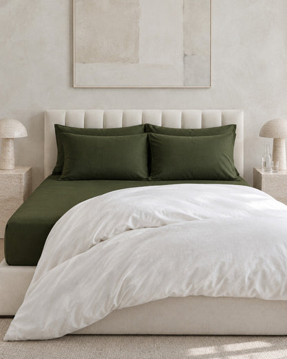 Forest green plain cotton fitted sheet against a limewashed plaster wall between travertine bedside tables