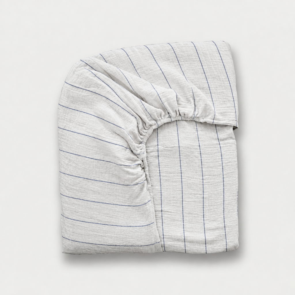 Folded cloud cotton fitted sheet with fine Blue Pinstripe on off-white crinkled muslin, plain background.