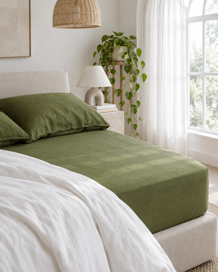 Moss green French linen fitted sheet beneath an arched window with a trailing pothos and rattan pendant