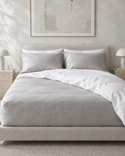 Pale silver-grey French linen fitted sheet against a limewashed plaster wall between travertine bedside tables
