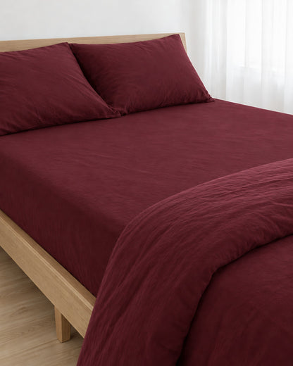 Burgundy Red plain cotton fitted sheet pulled taut over a mattress on an oak bed with matching pillows