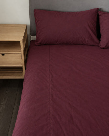Burgundy Red plain cotton fitted sheet and pillow on a bed beside an oak bedside table