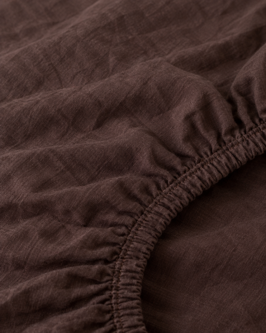 Macro of the gathered elastic corner of a Chocolate plain cotton fitted sheet.