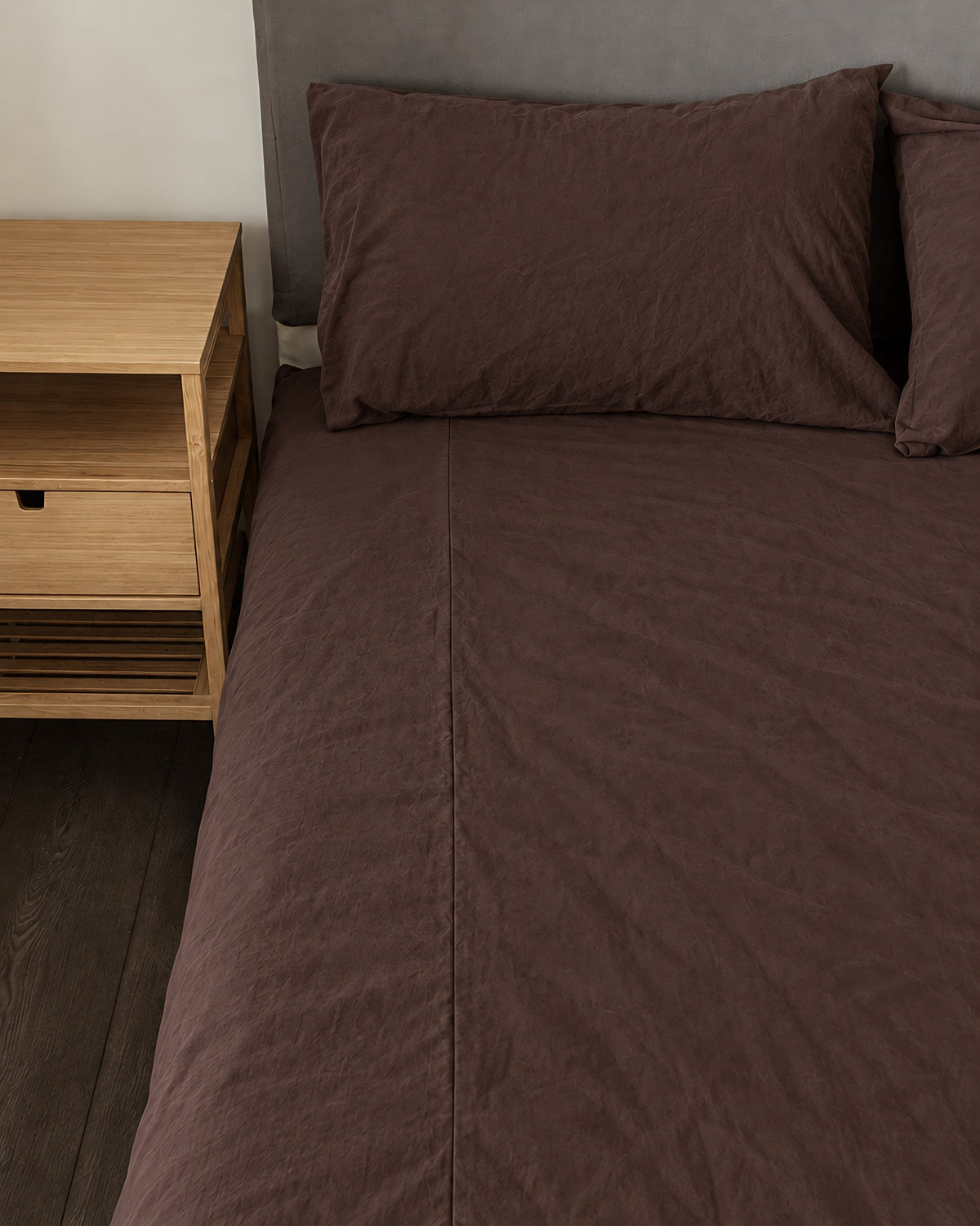 Chocolate plain cotton fitted sheet and pillow on a bed beside an oak bedside table