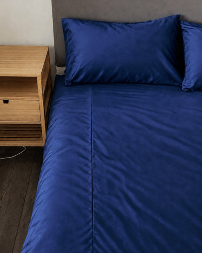 Marine Blue silky cotton fitted sheet, a navy blue cotton sateen fitted sheet, on a bed beside an oak bedside table