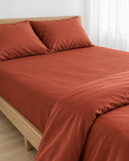 Rust silky cotton fitted sheet pulled taut over a mattress on an oak bed with matching pillows