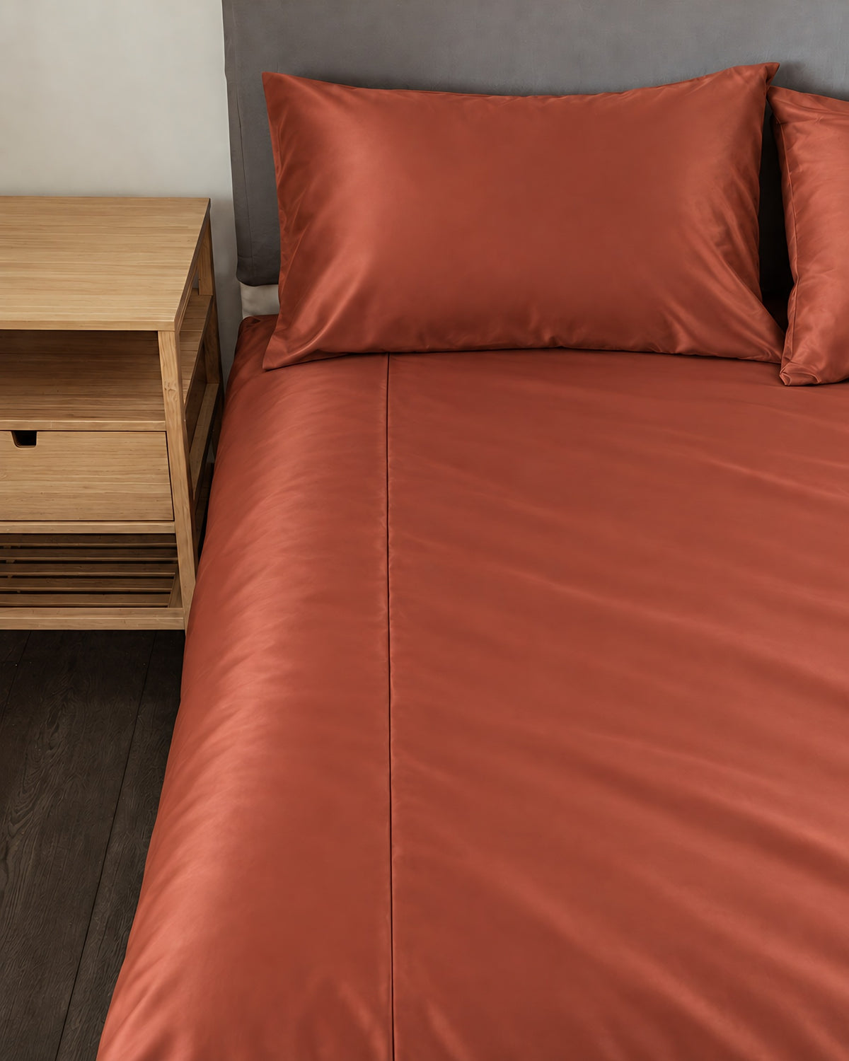 Rust silky cotton fitted sheet and pillow on a bed beside an oak bedside table