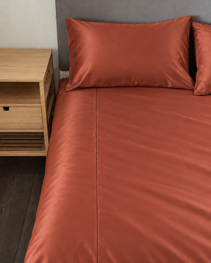 Rust silky cotton fitted sheet and pillow on a bed beside an oak bedside table