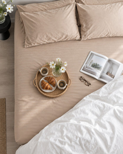 Beige silky cotton fitted sheet seen from above with a breakfast tray of coffee and croissants